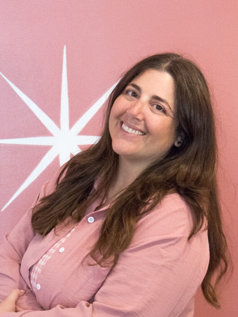 Headshot of Elise Kindya. She is wearing pink in front of the logo wall at PORTAL Therapy.