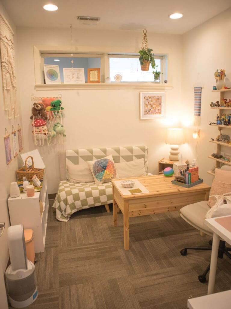 The inside of Elise Kindya's office at PORTAL Therapy. There is a small couch with green checkered blankets, a "Feelings Wheel" pillow, stuffed animals hanging on the wall, and lots of plants and light.