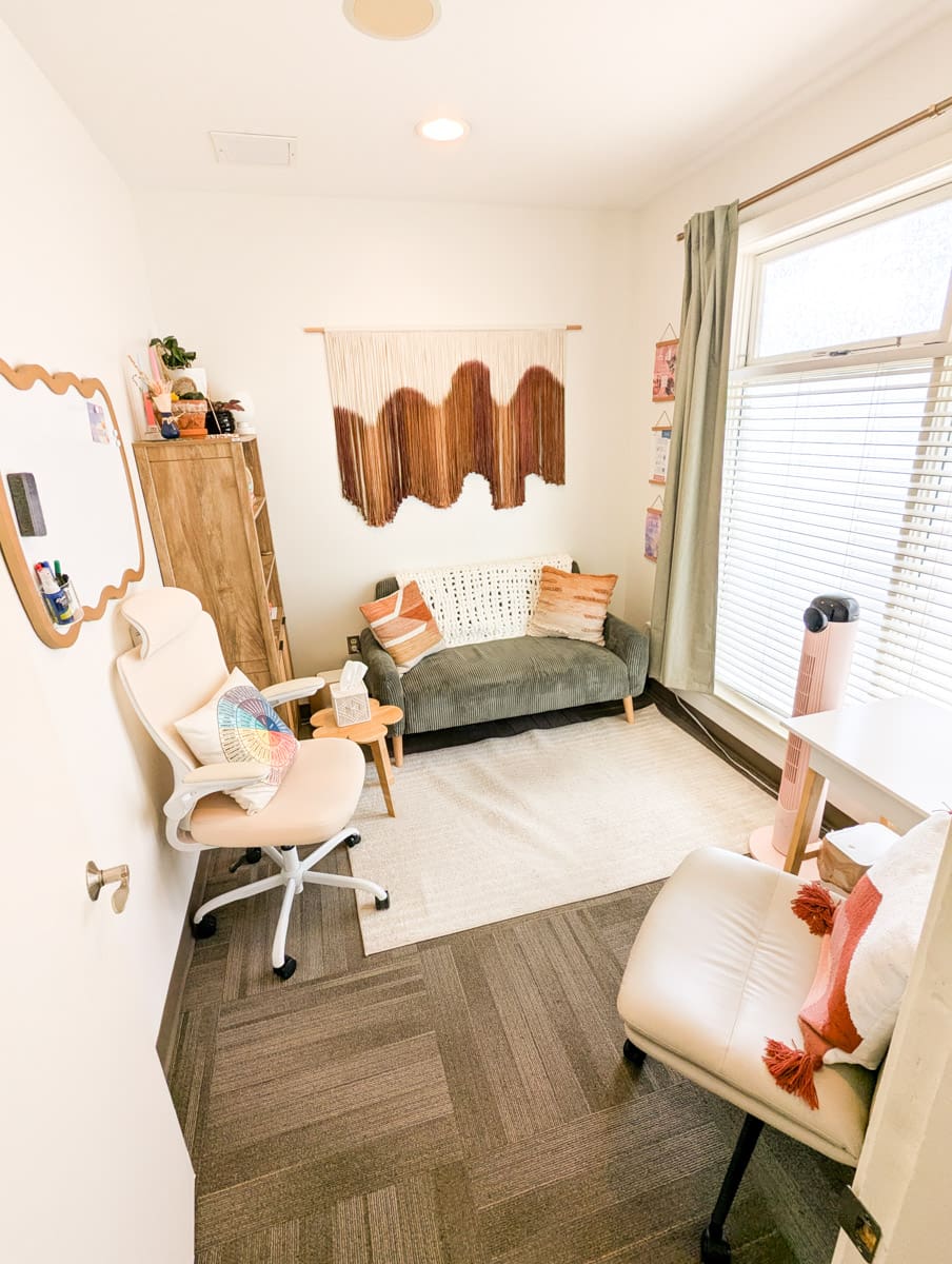 The interior of an office at PORTAL Therapy. There is a small couch with pillows, 2 desk chairs, a rug, a wall hanging and a lot of natural light from the large window.