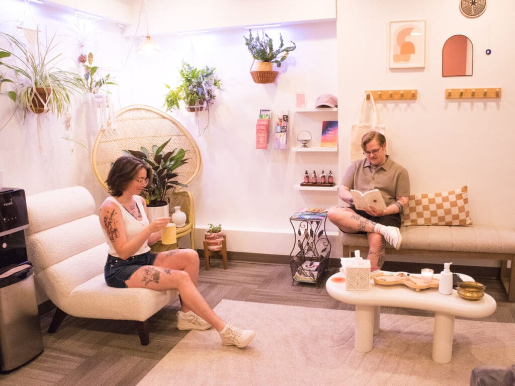 This photo shows two people in the PORTAL Therapy waiting room. They are both sitting. One is enjoying tea while the other is reading a book. There are plants and art in the photo as well.